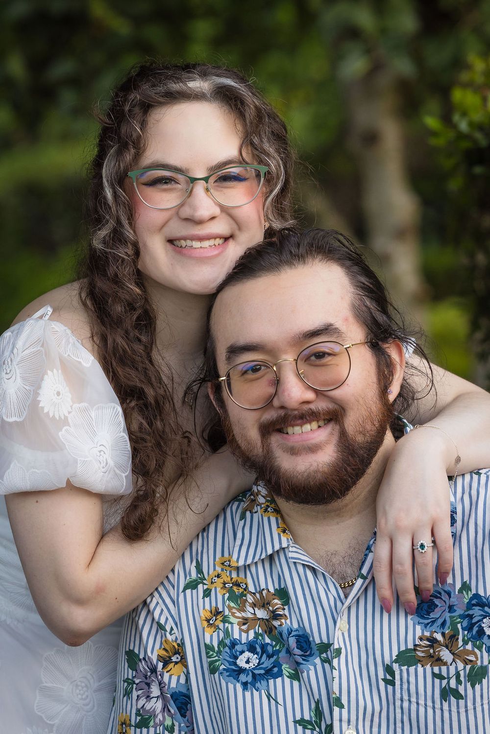 Smiling couple with glasses pose together outdoors, woman in white dress hugging man in floral shirt.