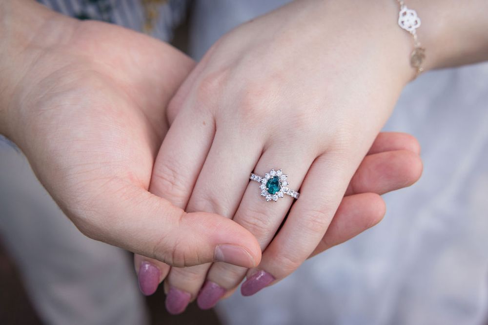 Close-up of hands showing an elegant oval emerald and diamond halo engagement ring on a woman's finger.