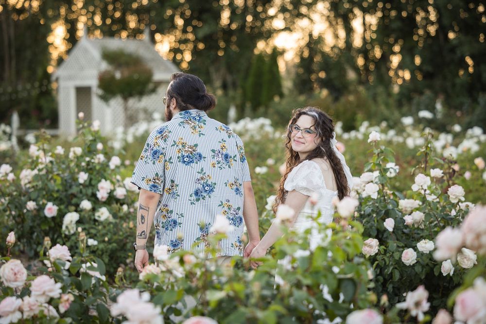 Couple holding hands in a romantic rose garden at golden hour, surrounded by blooming pink and white roses.