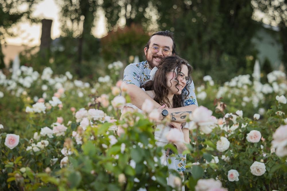 Couple embracing and smiling in a lush rose garden with white blooms at golden hour.