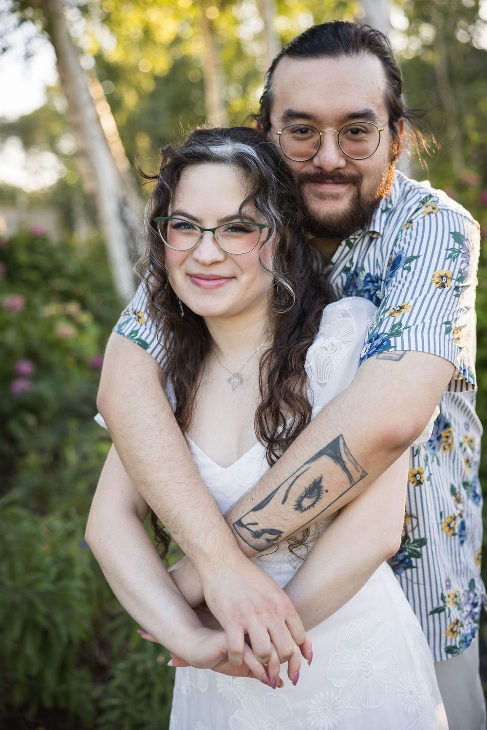 Couple embracing outdoors, man with tattoos in floral shirt hugs woman in white dress among garden greenery.
