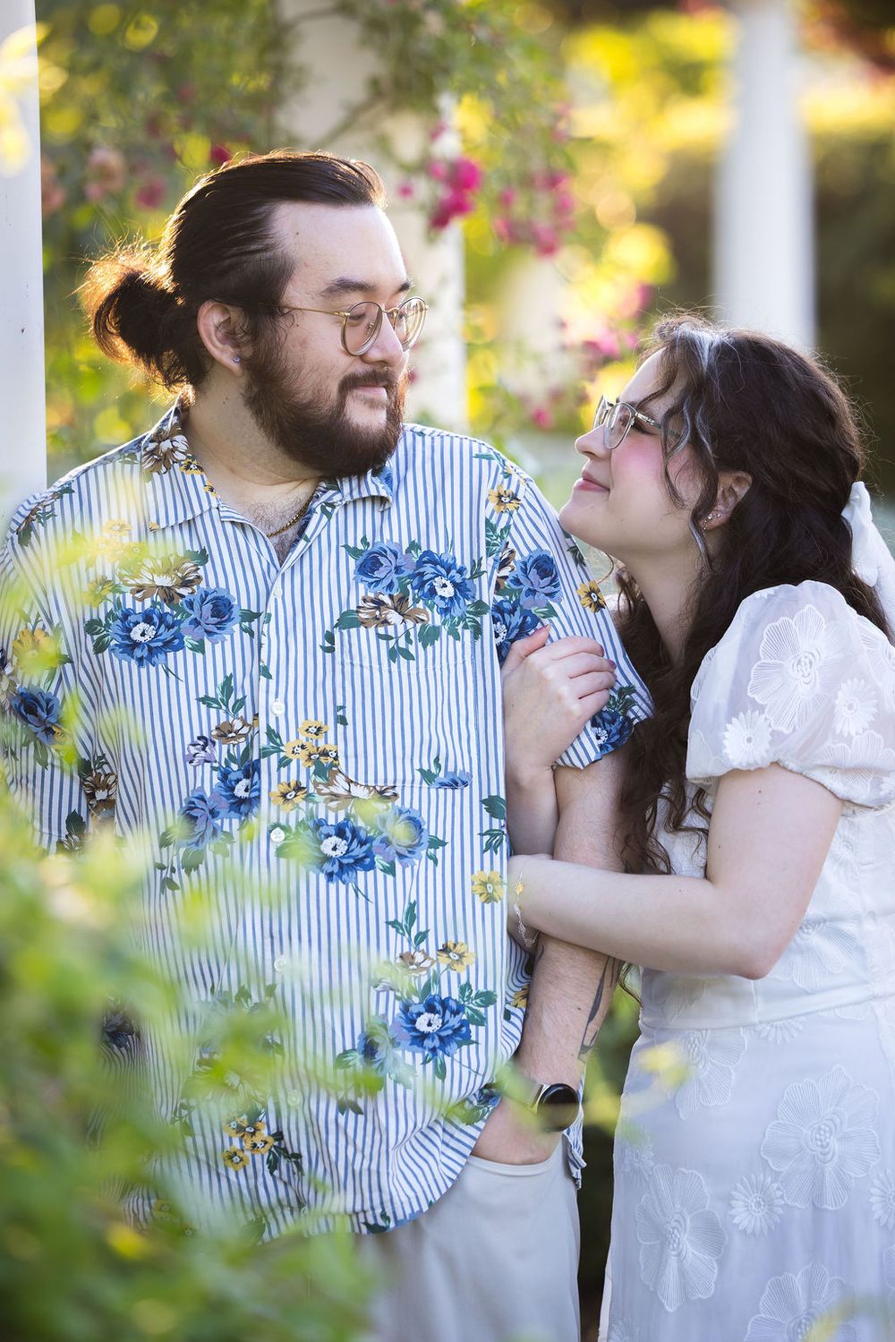 A couple in floral and white outfits share a tender moment outdoors surrounded by lush greenery and blooming flowers.
