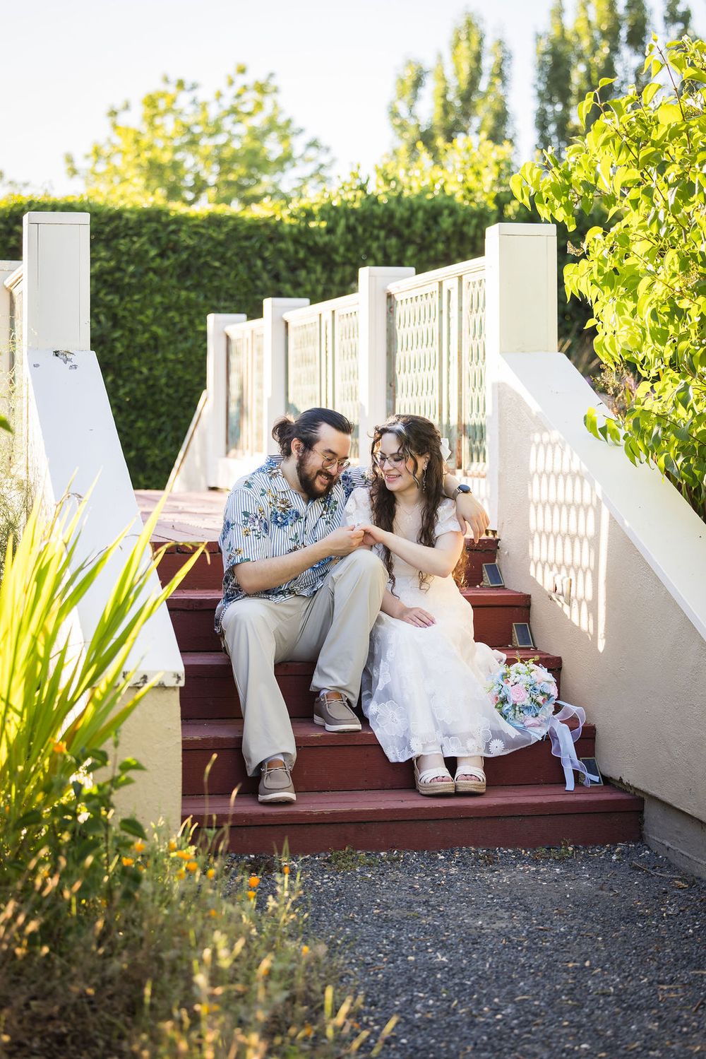 Happy couple in wedding attire sitting on red outdoor steps holding hands surrounded by lush greenery.