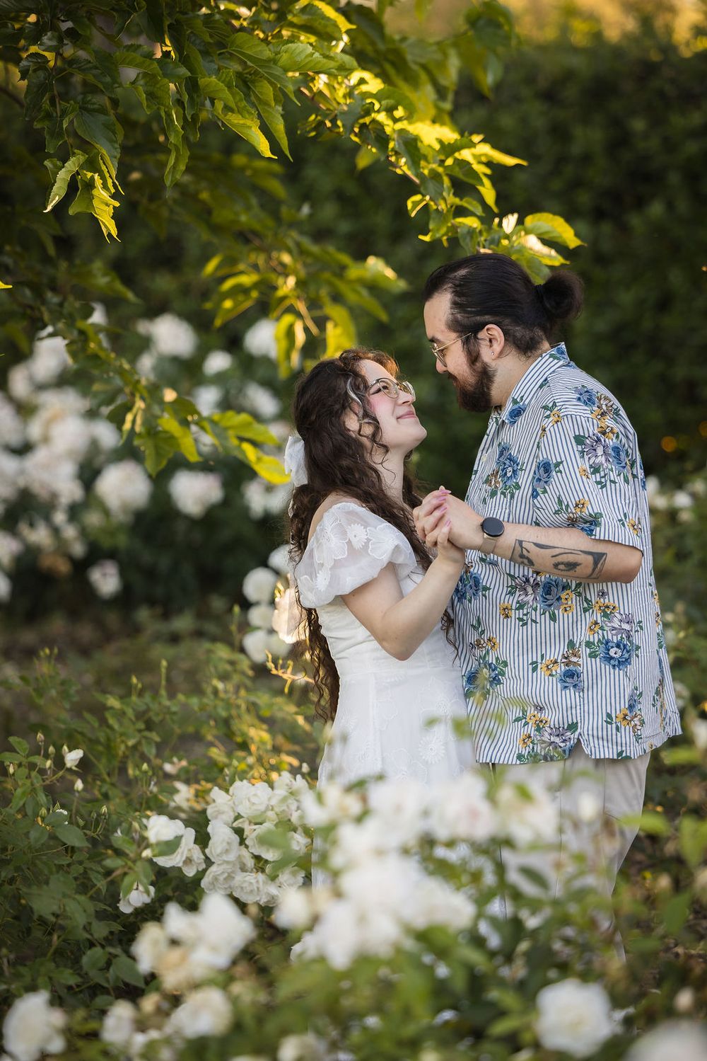 A couple holds hands and gazes lovingly at each other among white roses in a lush garden.