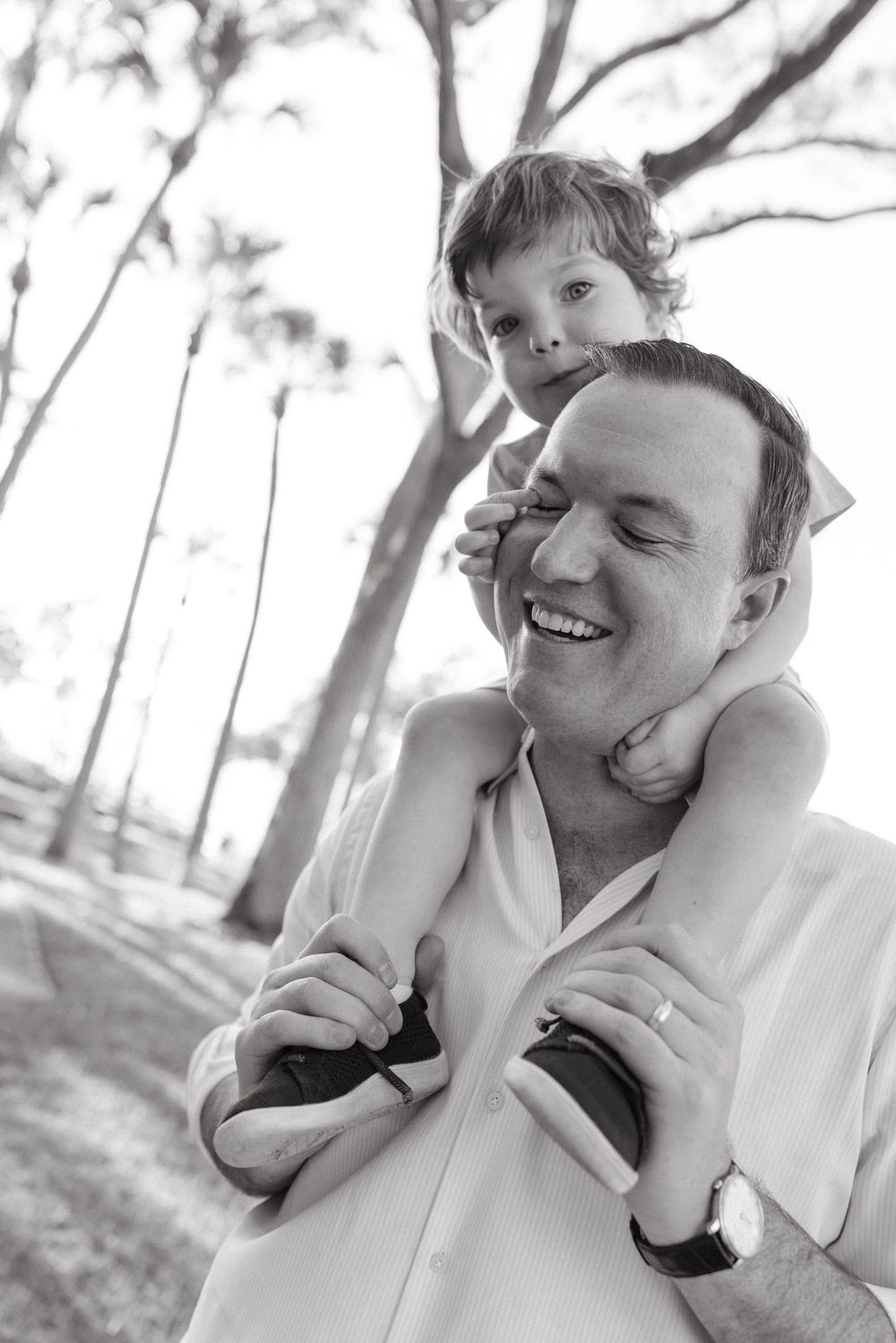 Black and white photo of a laughing father carrying his young child on his shoulders in a park.