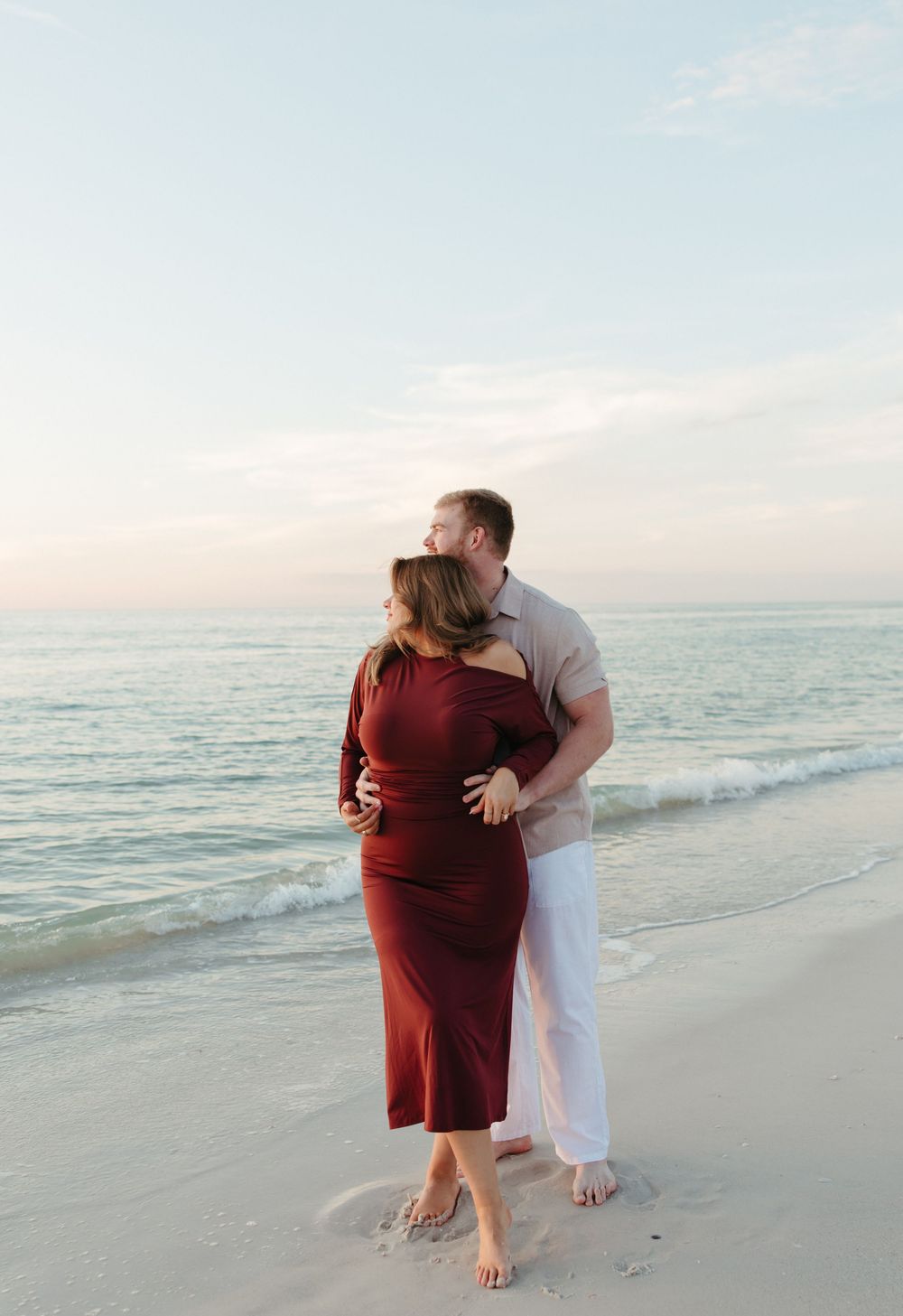 Couple embracing on a sandy beach at sunset, woman in red dress, man in white pants, ocean waves behind them.