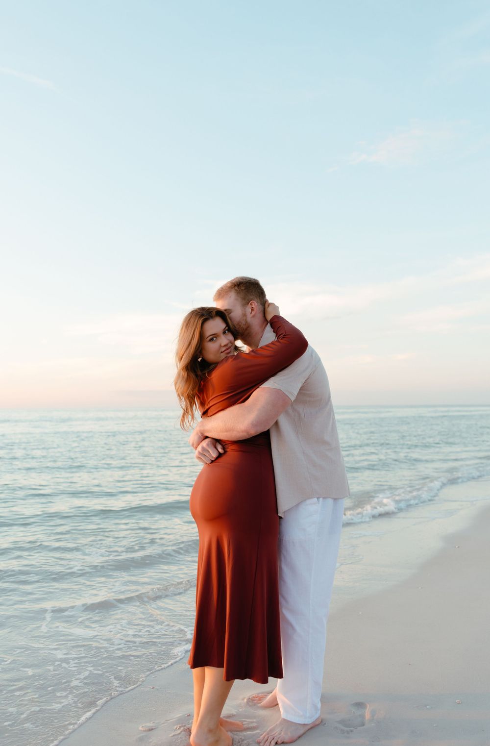 Pregnant couple embracing on a beach at sunset, woman in rust dress, man in white linen outfit.