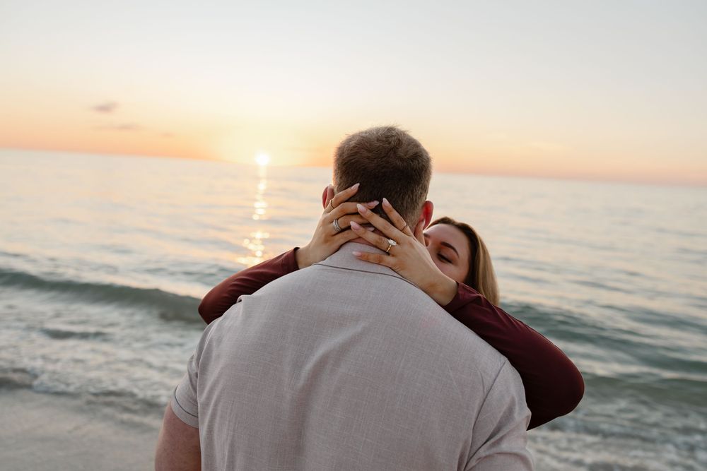 Couple embracing at sunset on the beach, woman showing engagement ring while ocean waves shimmer in golden light.