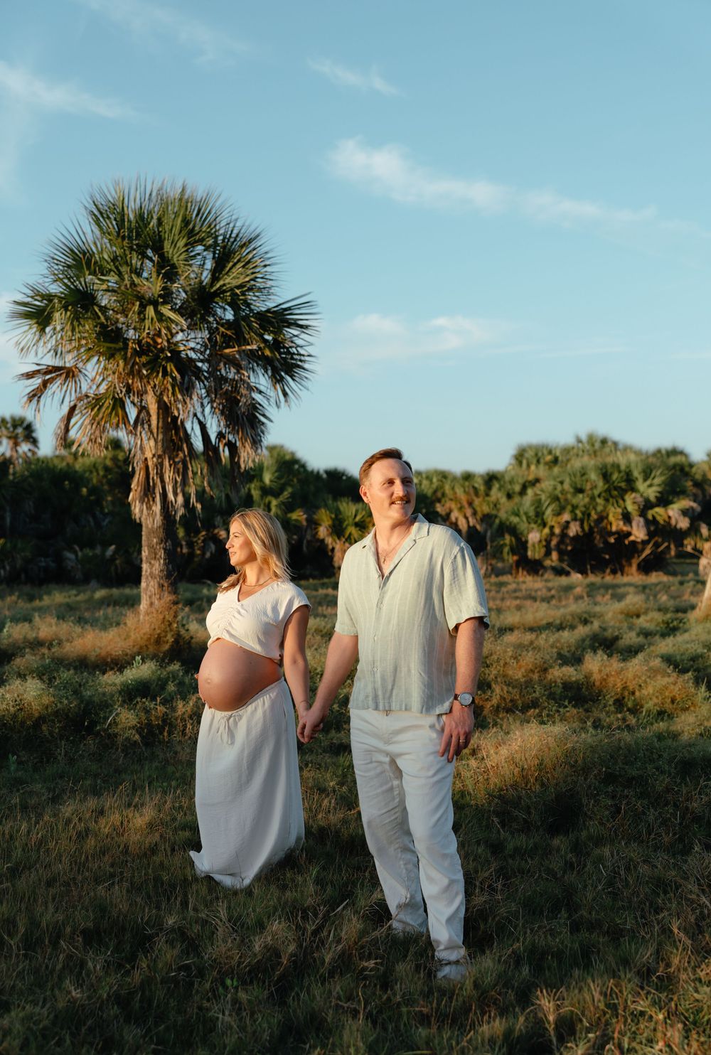 Pregnant couple holding hands in a sunny field with palm trees, wearing white outfits during a maternity photo session.