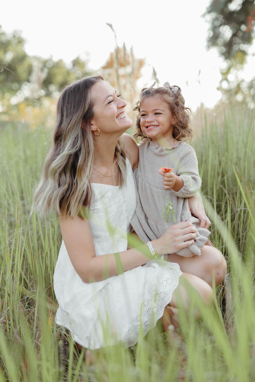 A smiling woman in a white dress holds a happy toddler girl in a green field during golden hour.