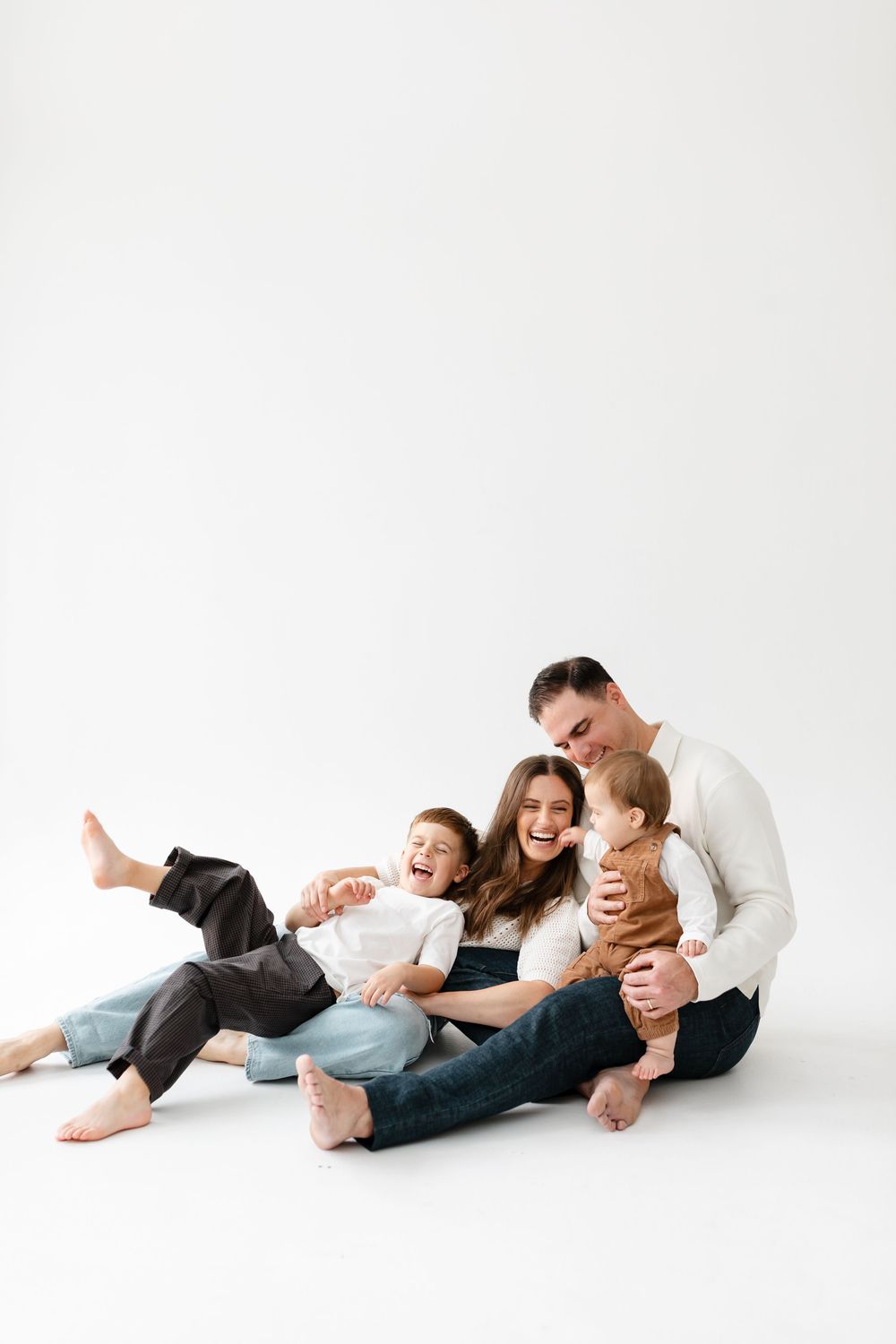 Happy family of four sitting on white background, parents laughing with young children in casual white and denim outfits.