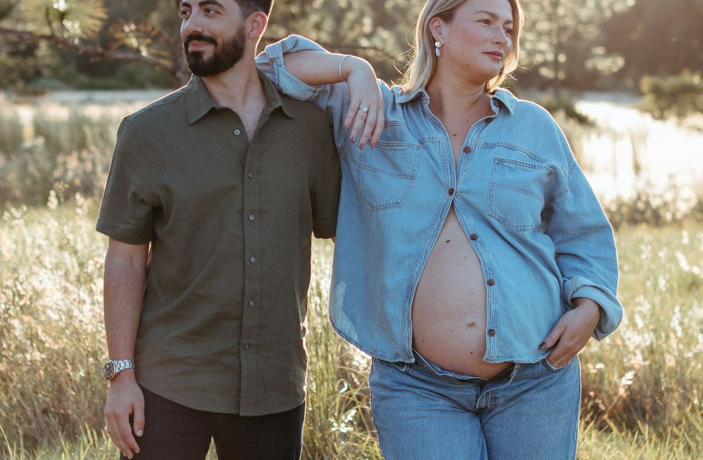 Couple posing outdoors in a sunny field, woman showing pregnant belly in denim shirt, man in olive shirt beside her.