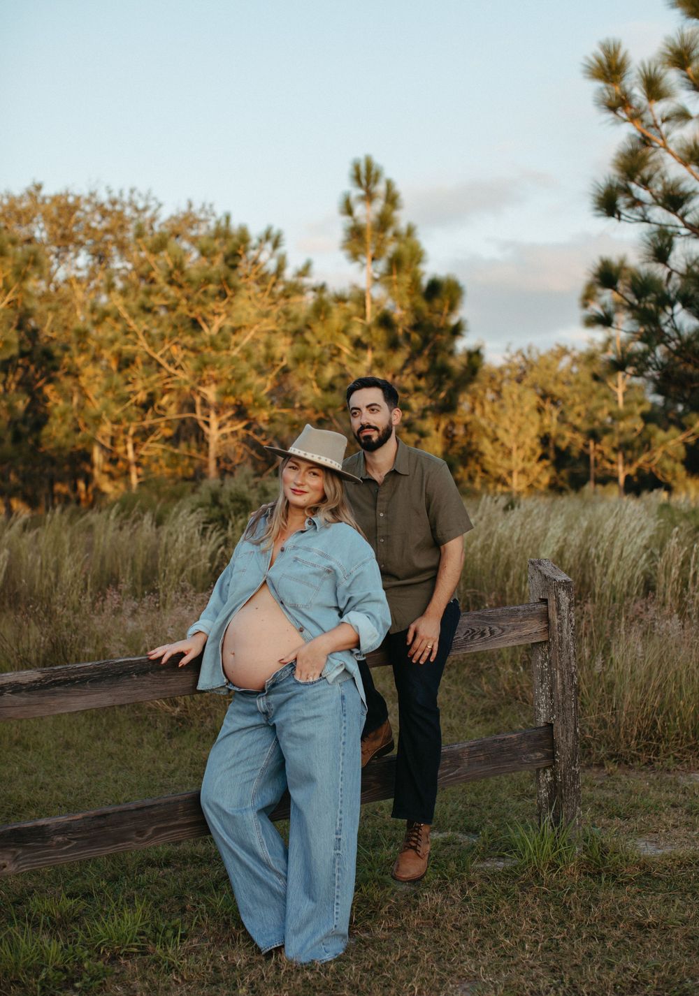 Pregnant woman in denim outfit and hat poses with bearded man by rustic fence at golden hour outdoors.