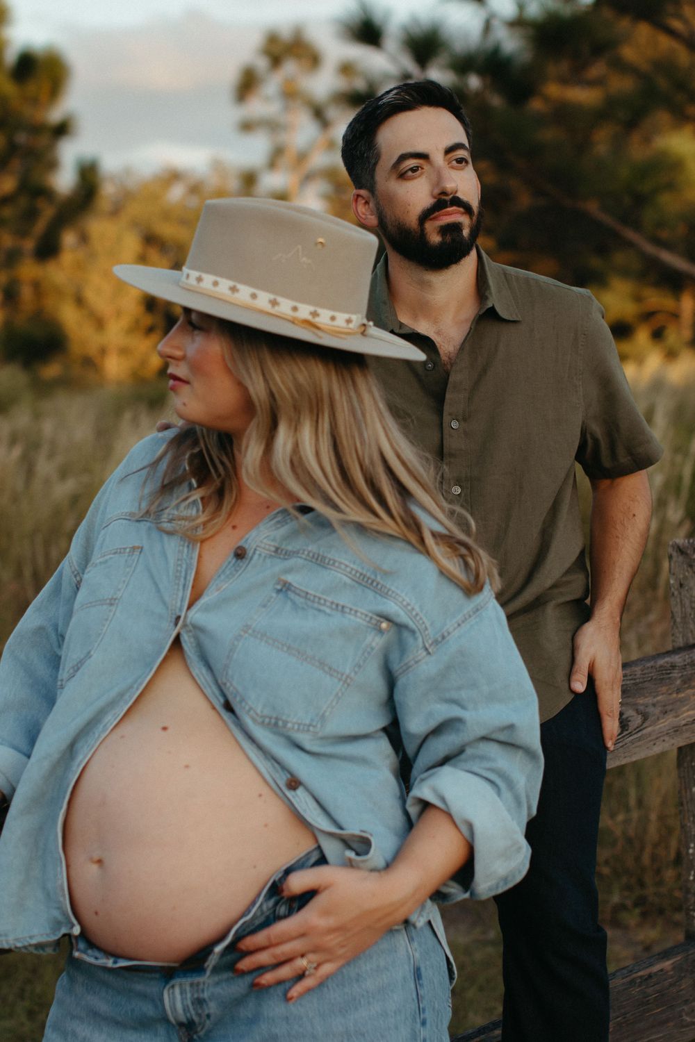 Pregnant woman in denim jacket and wide-brim hat poses outdoors with bearded man during golden hour maternity shoot.