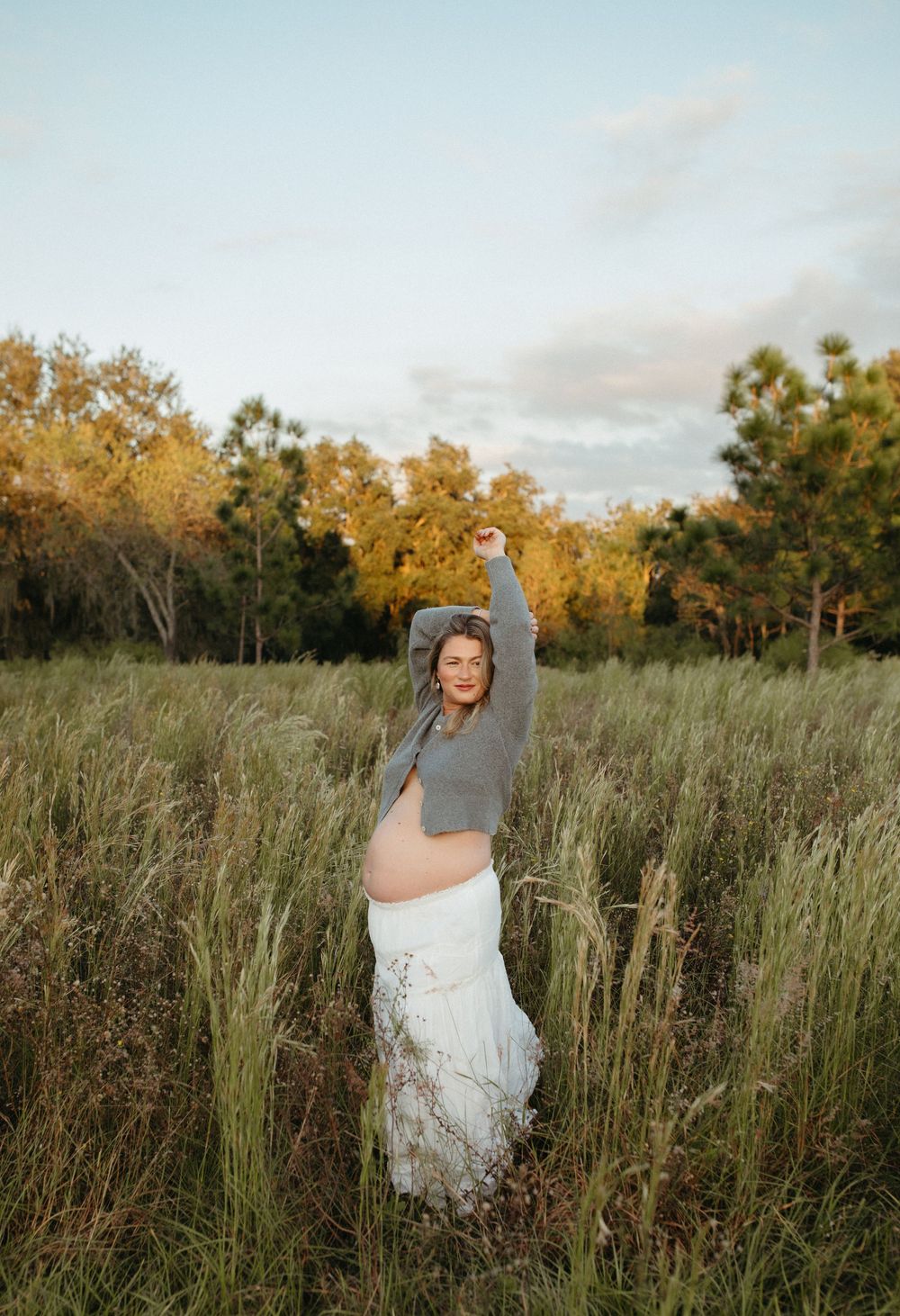 Pregnant woman in gray crop top and white skirt posing in a green field with arms raised at golden hour.