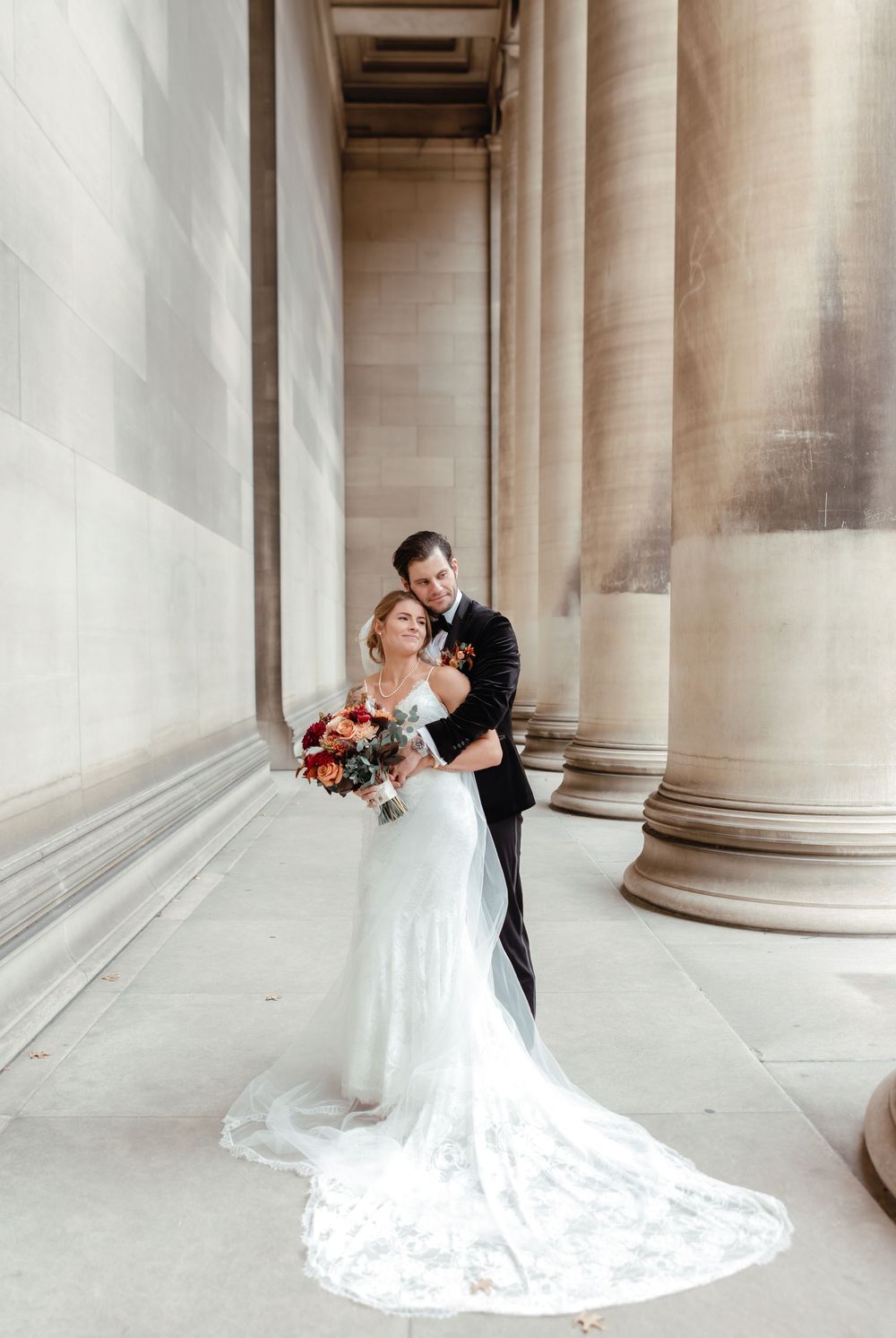 Bride and groom embrace among grand stone columns, bride wearing lace gown with long train holding colorful bouquet.