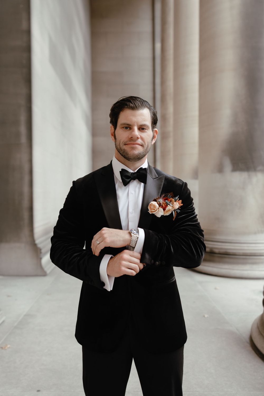 Dapper groom in black velvet tuxedo with bow tie and floral boutonniere adjusting cufflinks outdoors.
