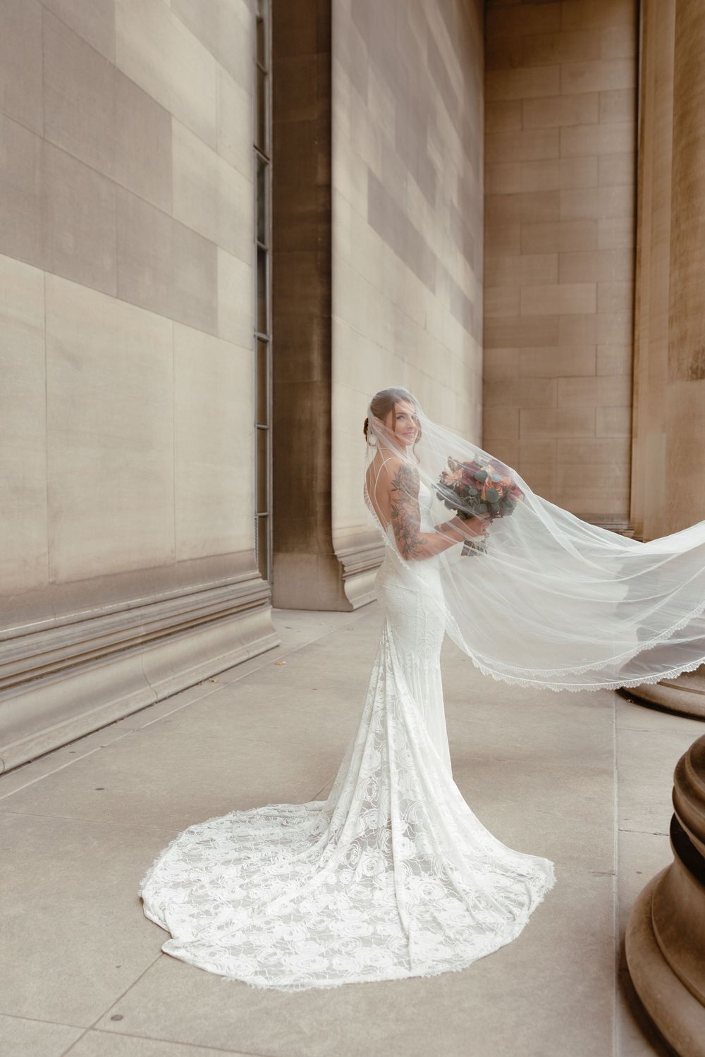 Bride in lace mermaid gown with flowing veil billowing beside stone columns, holding floral bouquet.