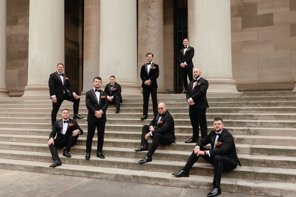 Groomsmen in black tuxedos posing on stone steps of a grand building for a wedding photo.