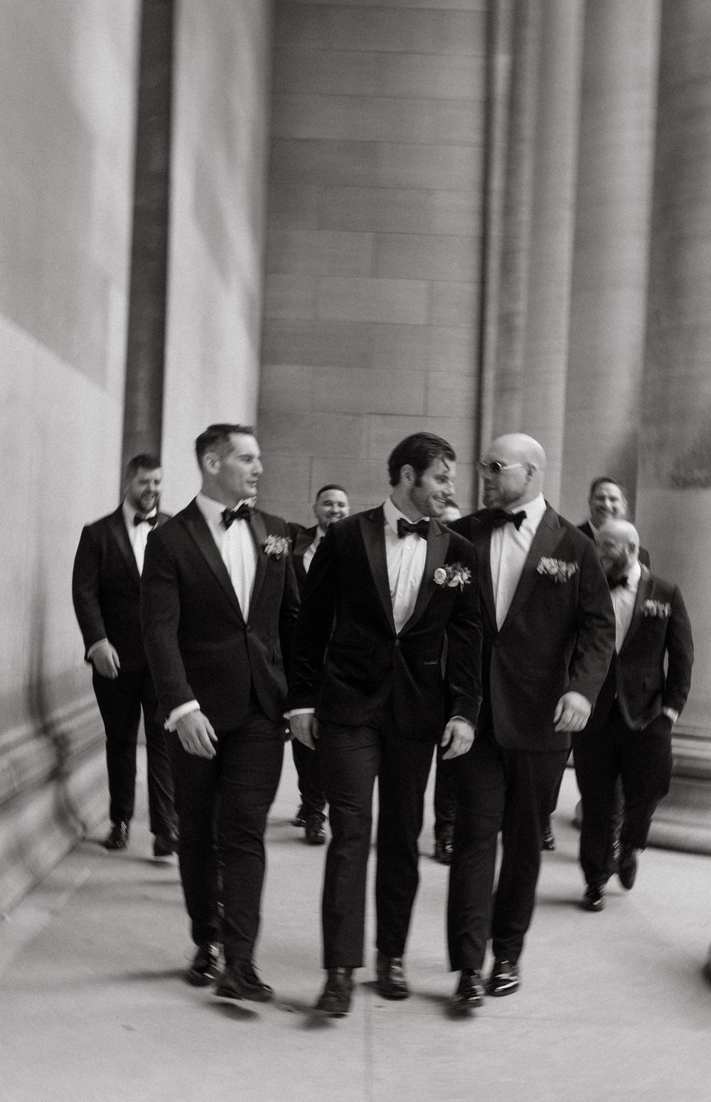 Black and white photo of groomsmen in tuxedos walking together down a grand hallway with boutonnieres.