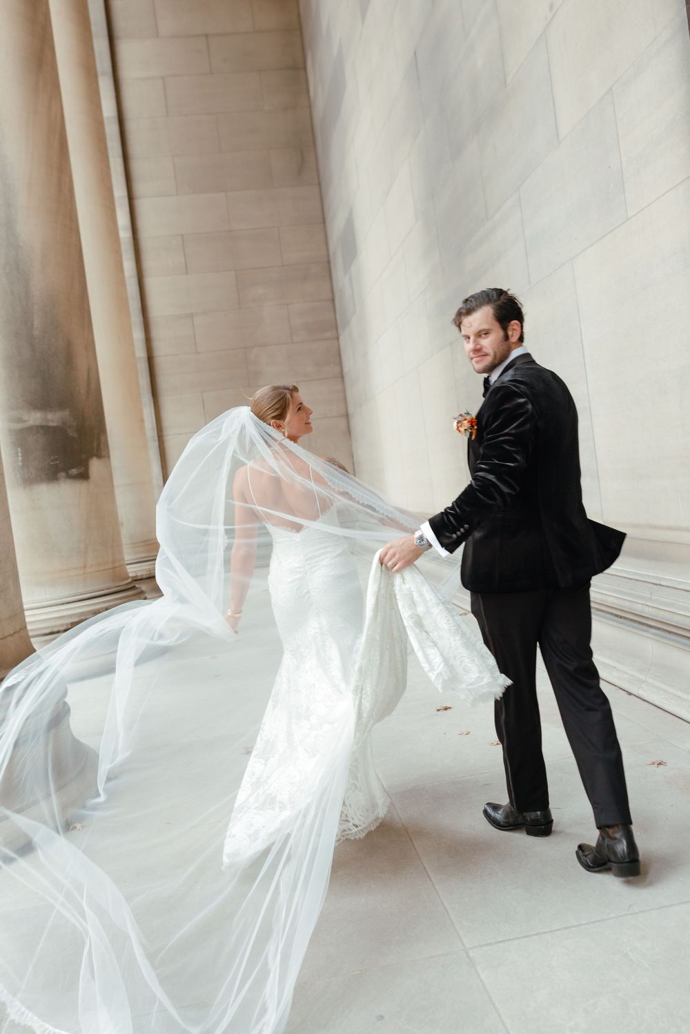 Bride in flowing white gown and veil walks with groom in black tuxedo near stone columns.
