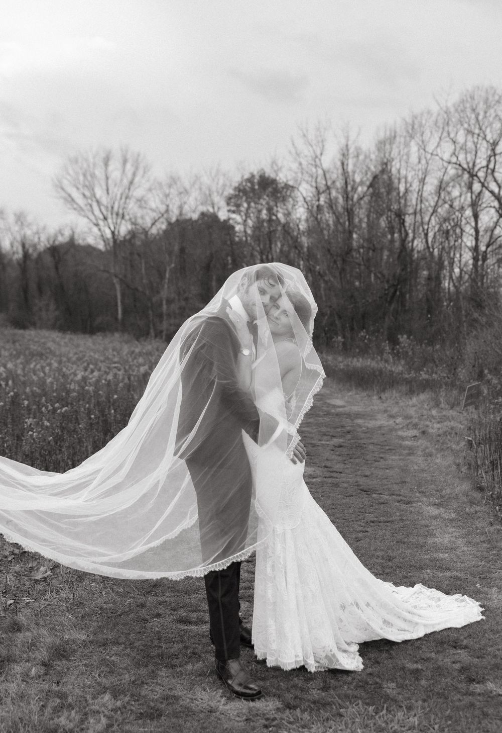 Bride and groom sharing a romantic kiss under a flowing veil in a rustic outdoor setting.