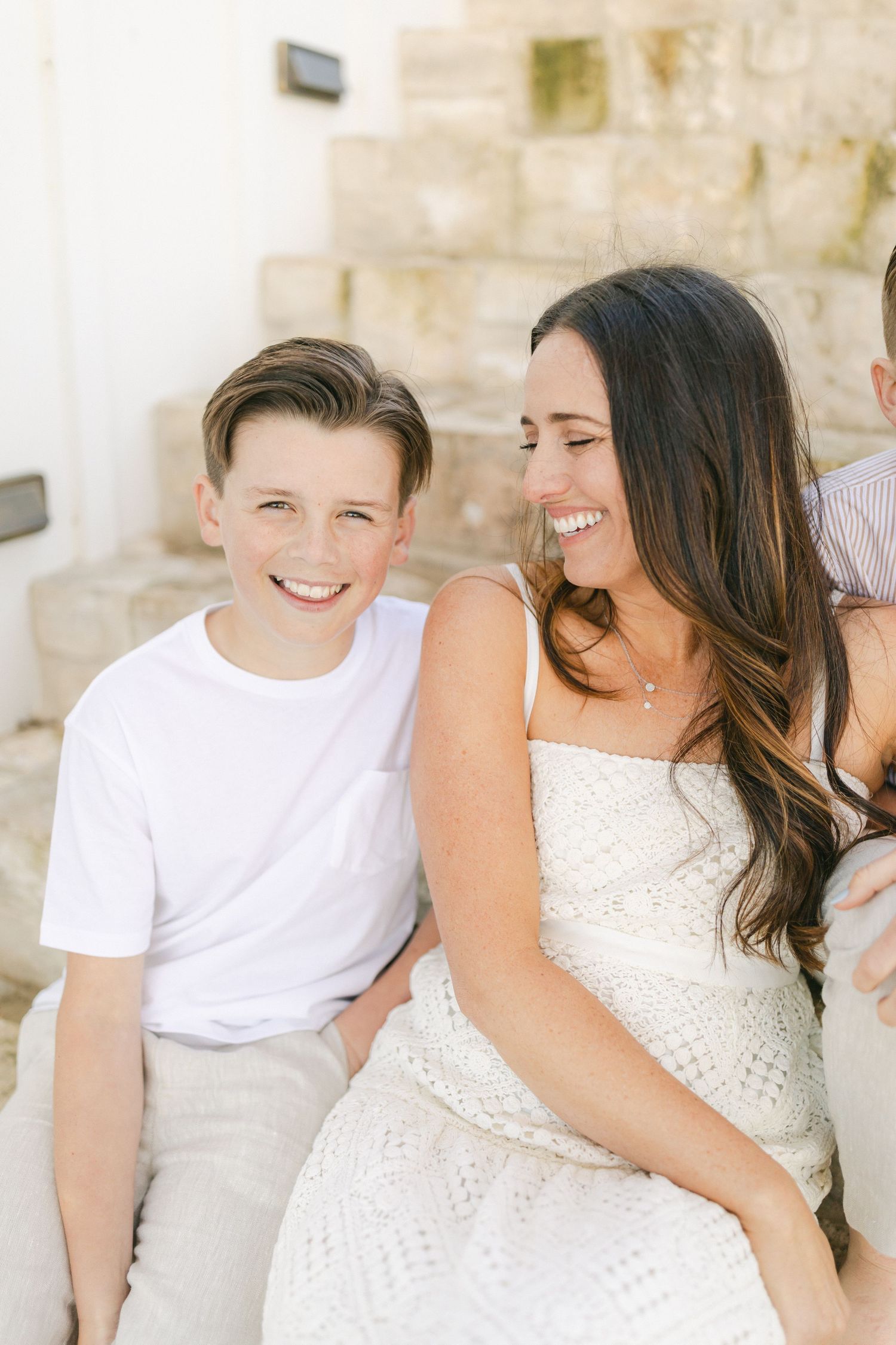 A smiling boy in a white t-shirt sits beside a laughing brunette woman in a white dress on outdoor steps.
