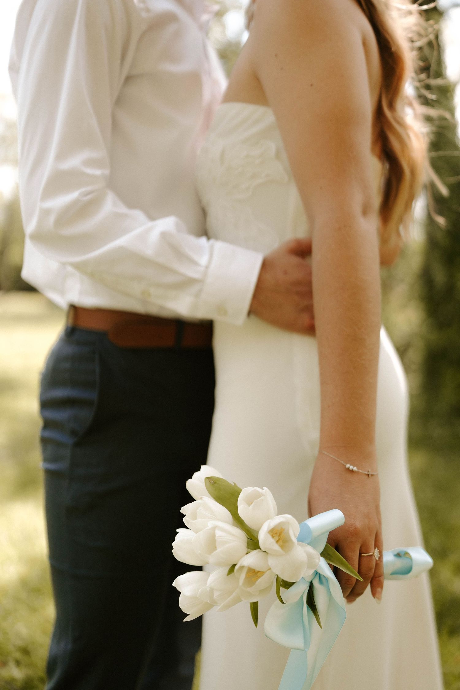 Bride holding white tulip bouquet with blue ribbon stands beside groom in white shirt and navy pants outdoors.
