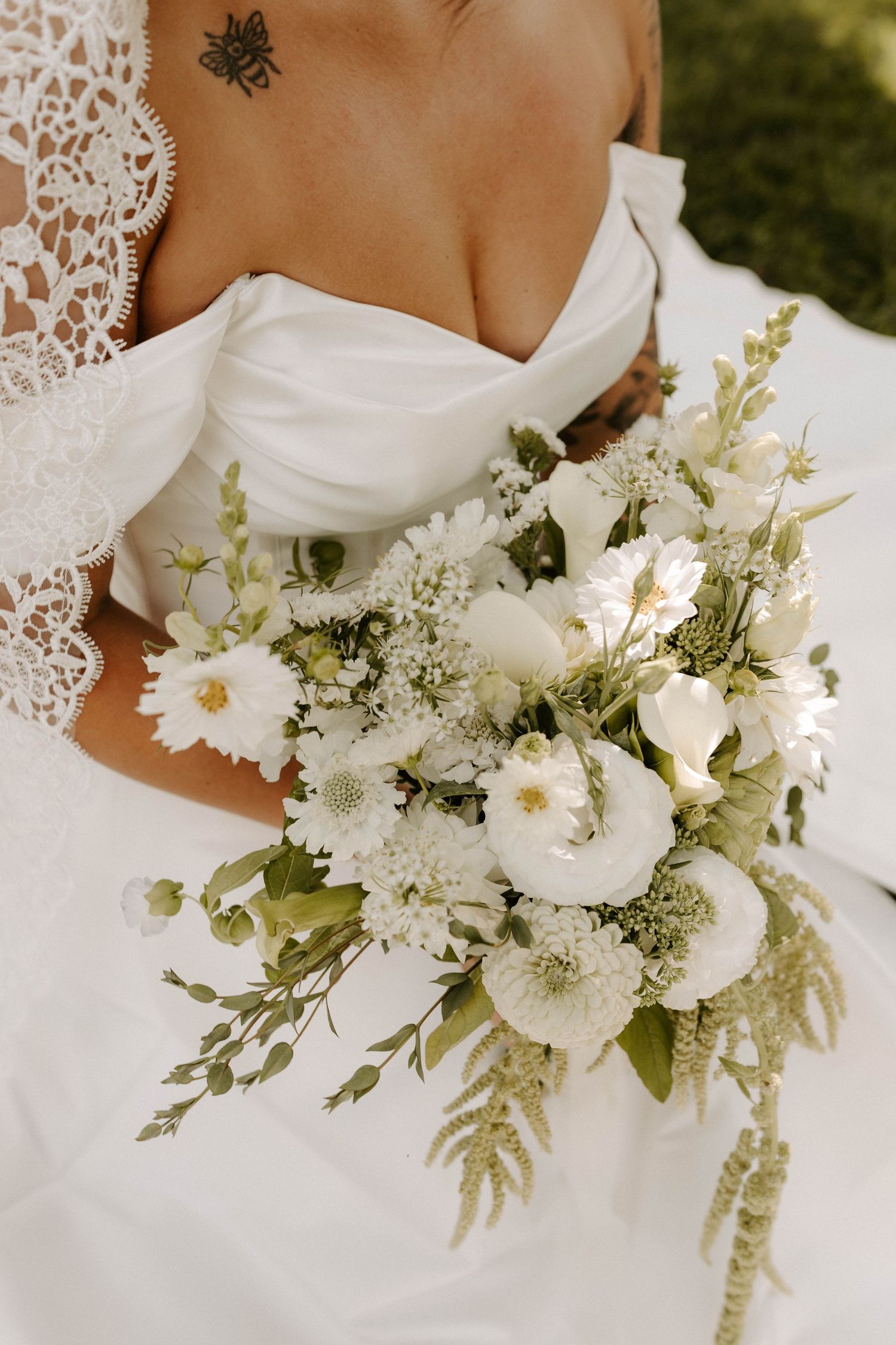 Bride holding white tulip bouquet with blue ribbon stands beside groom in white shirt and navy pants outdoors.