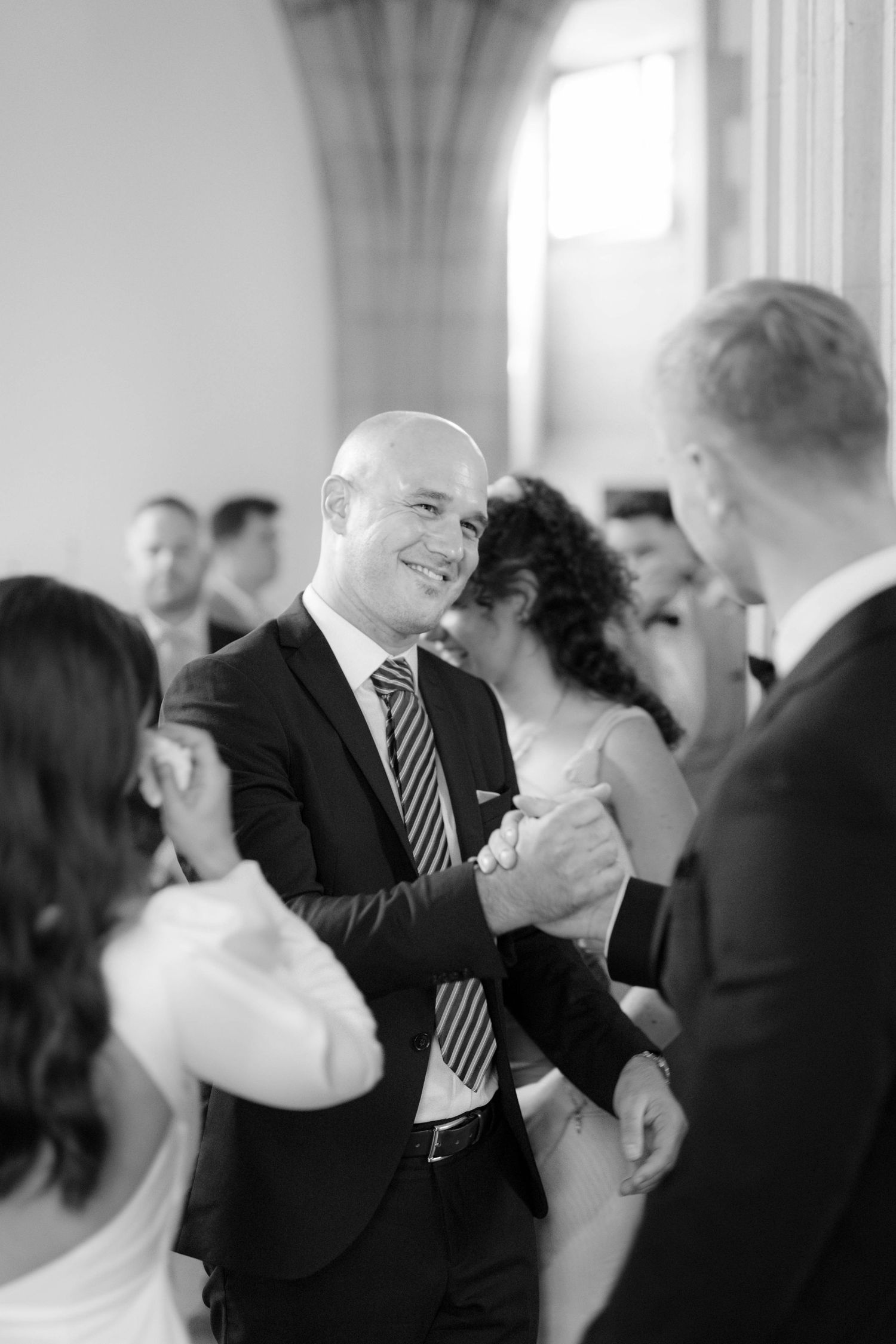 Bald man in suit smiling warmly while shaking hands at a formal wedding reception in black and white.