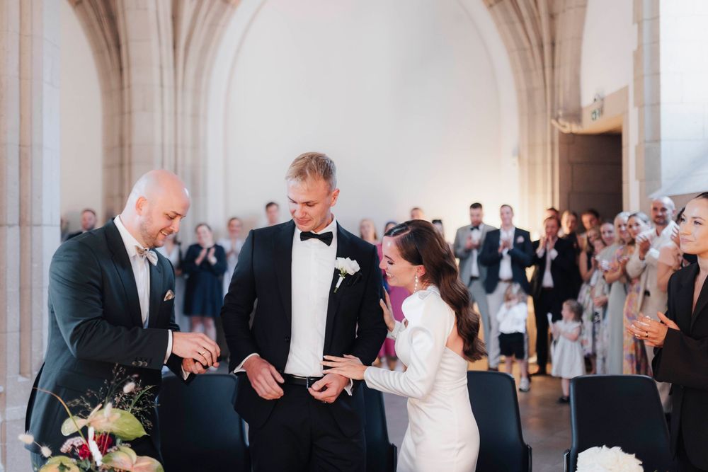 Bride and groom exchange rings at elegant wedding ceremony while officiant assists, guests applauding in bright venue.