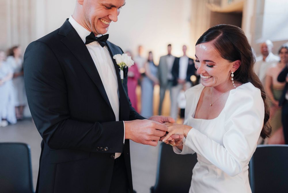Groom placing wedding ring on bride's finger during ceremony, both smiling, guests visible in background.