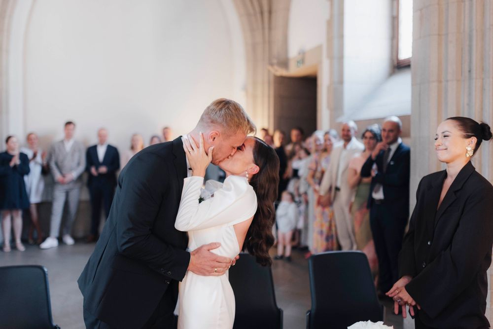 Bride and groom share first kiss at wedding ceremony while guests watch and smiling woman stands nearby.
