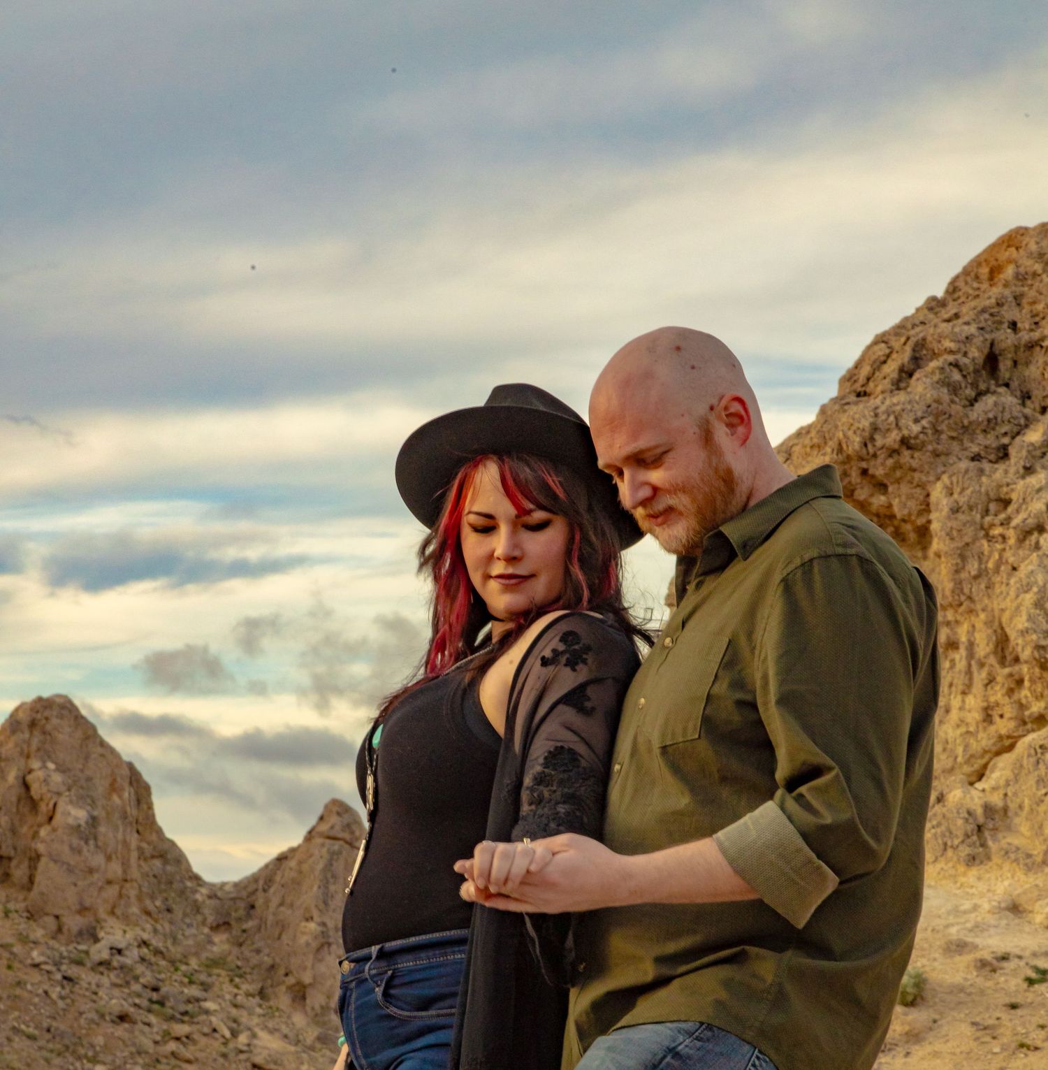 A couple poses romantically in a desert landscape, woman with red hair and black hat, man in olive shirt.
