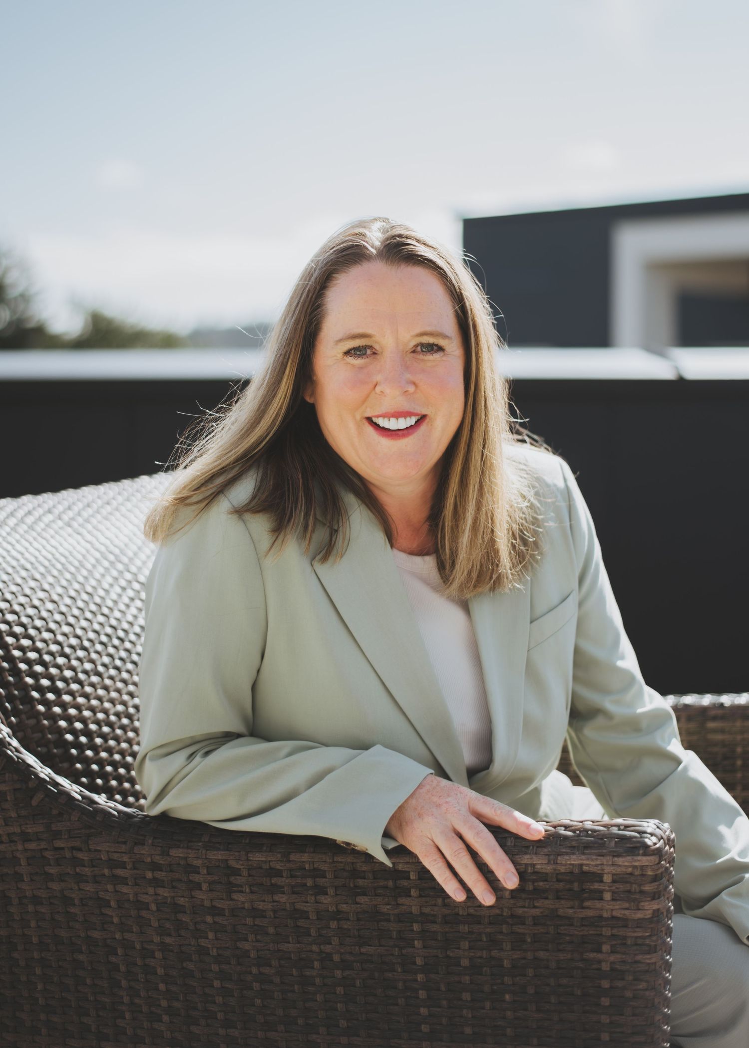Professional woman in light green blazer smiling outdoors, seated in wicker chair on sunny patio.