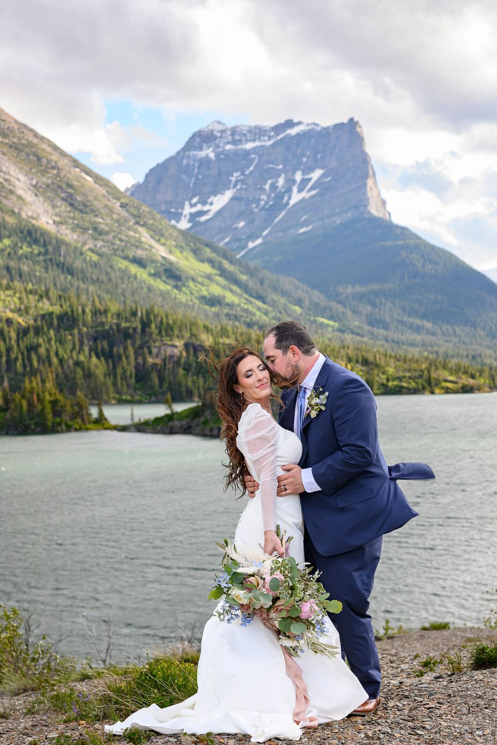 Bride and groom embrace near a mountain lake, she holds a floral bouquet in a white gown, he wears a navy suit.