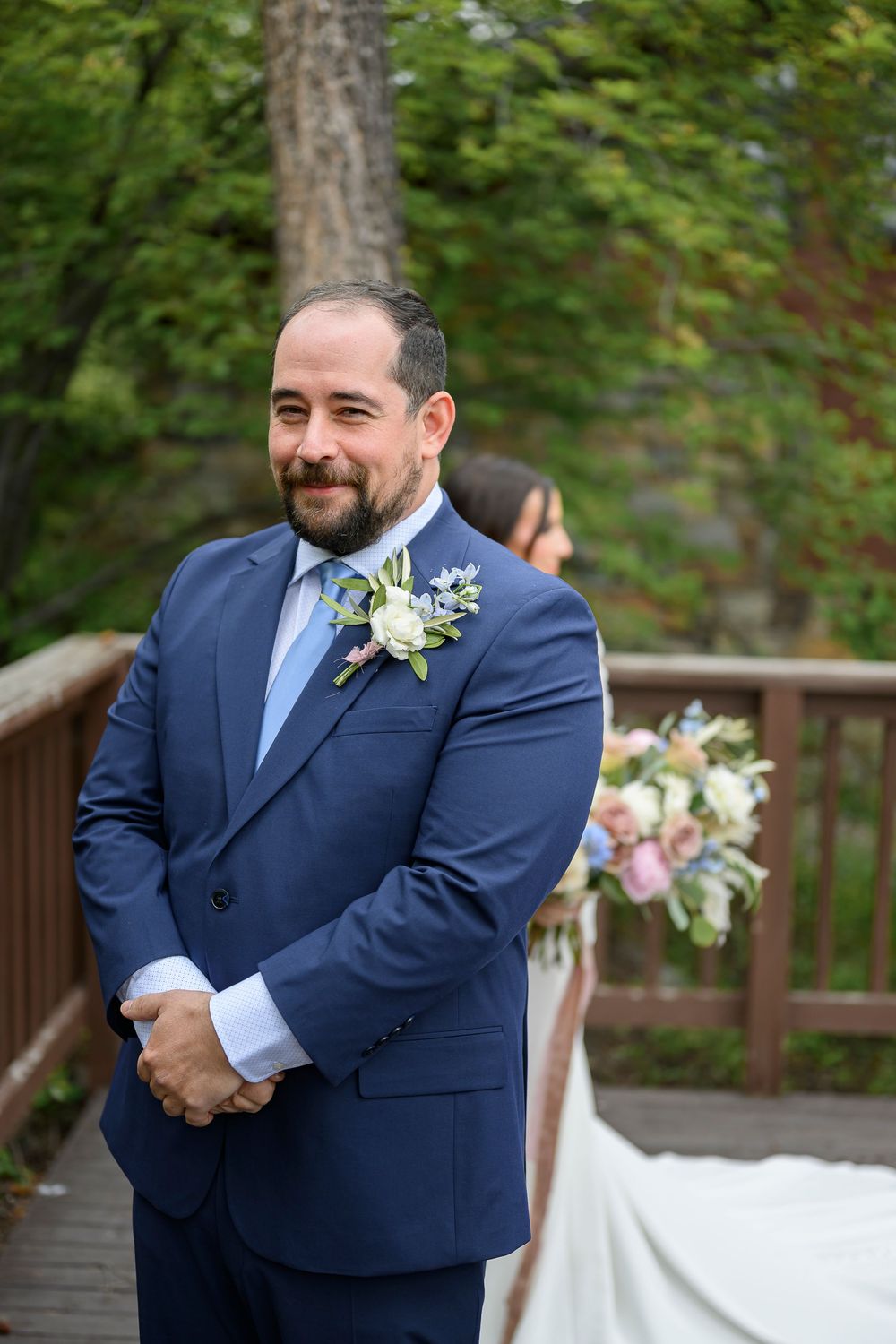 Groom in blue suit with boutonniere smiling on wooden bridge, bride with floral bouquet visible in background.