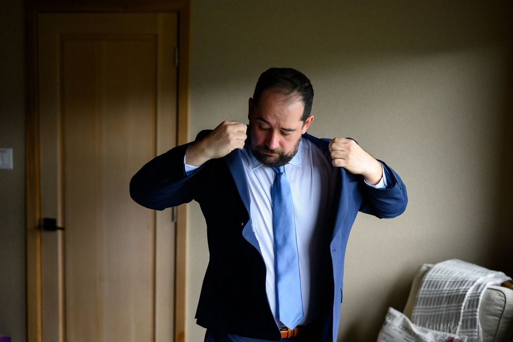 Man in blue suit adjusting jacket collar while getting dressed in a room.