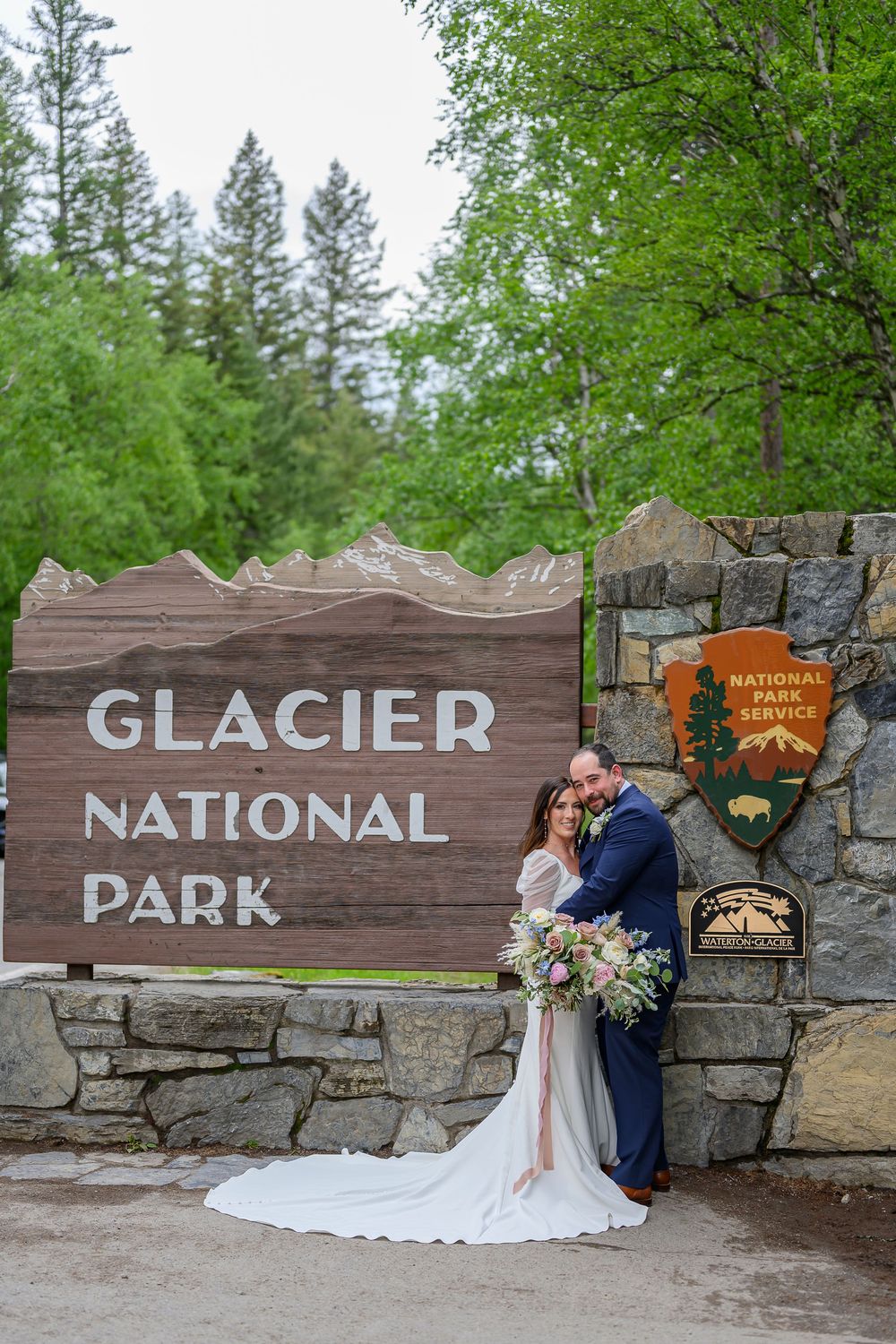 Couple in wedding attire poses in front of Glacier National Park entrance sign surrounded by lush green trees.