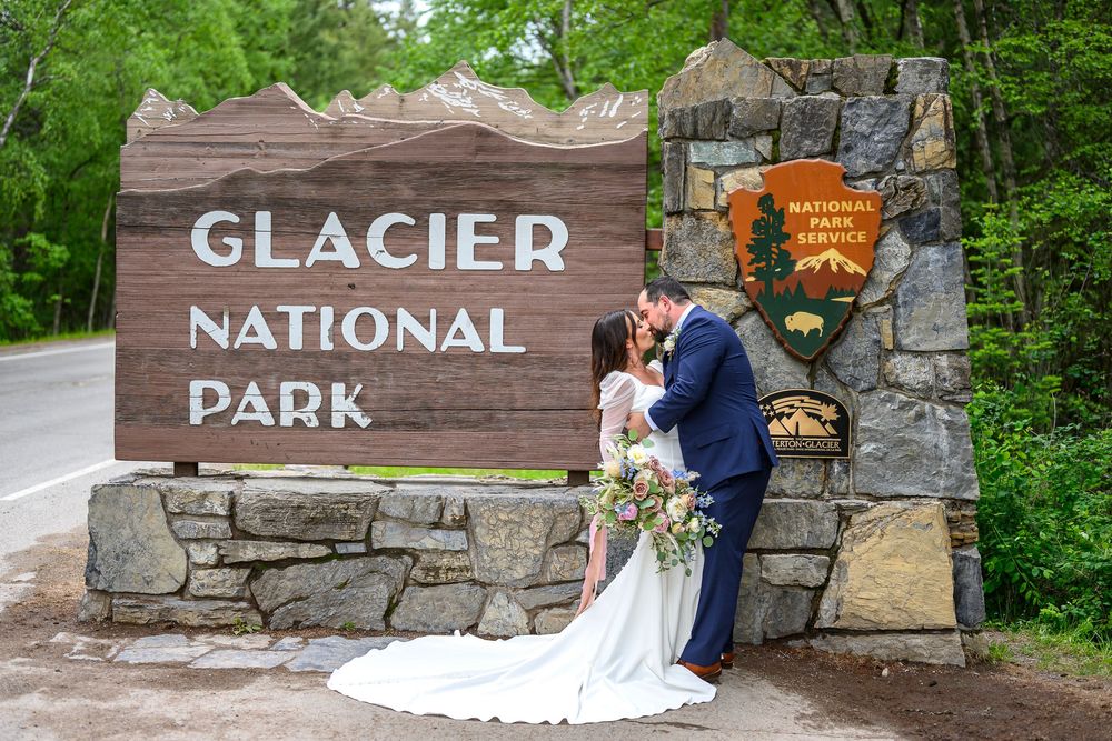 Couple kissing in wedding attire in front of Glacier National Park entrance sign surrounded by lush green trees.