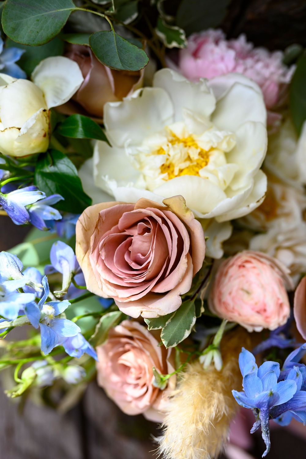 Elegant bouquet featuring dusty pink roses, white peonies, and blue delphinium flowers arranged together.