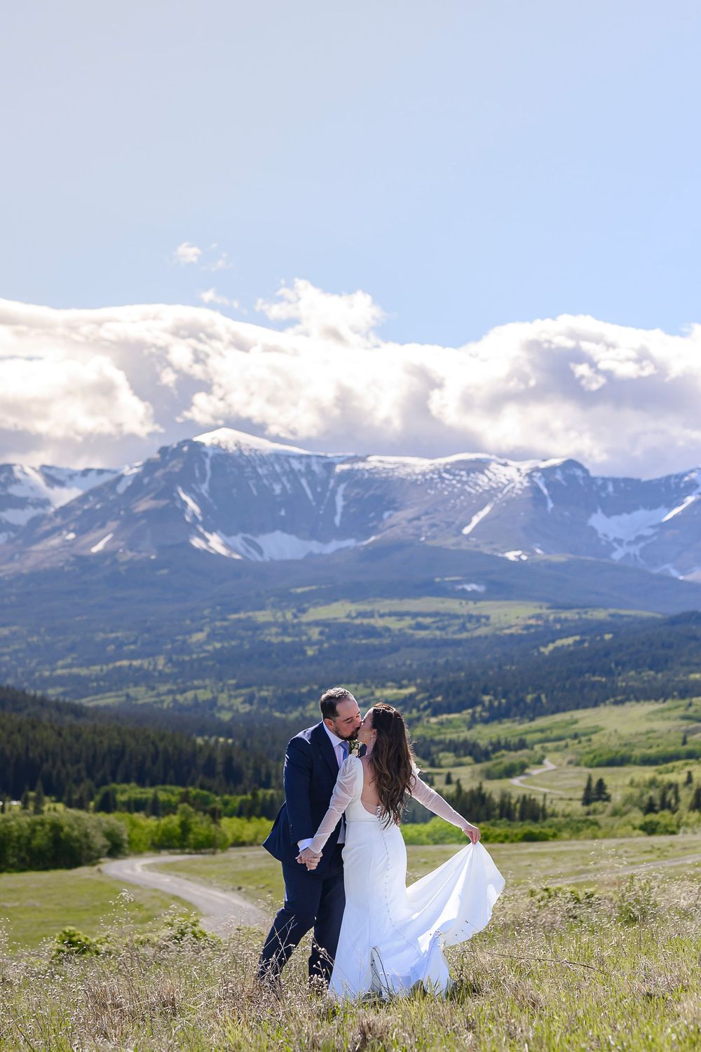 Bride and groom kissing in a mountain meadow with snow-capped peaks and blue sky backdrop.