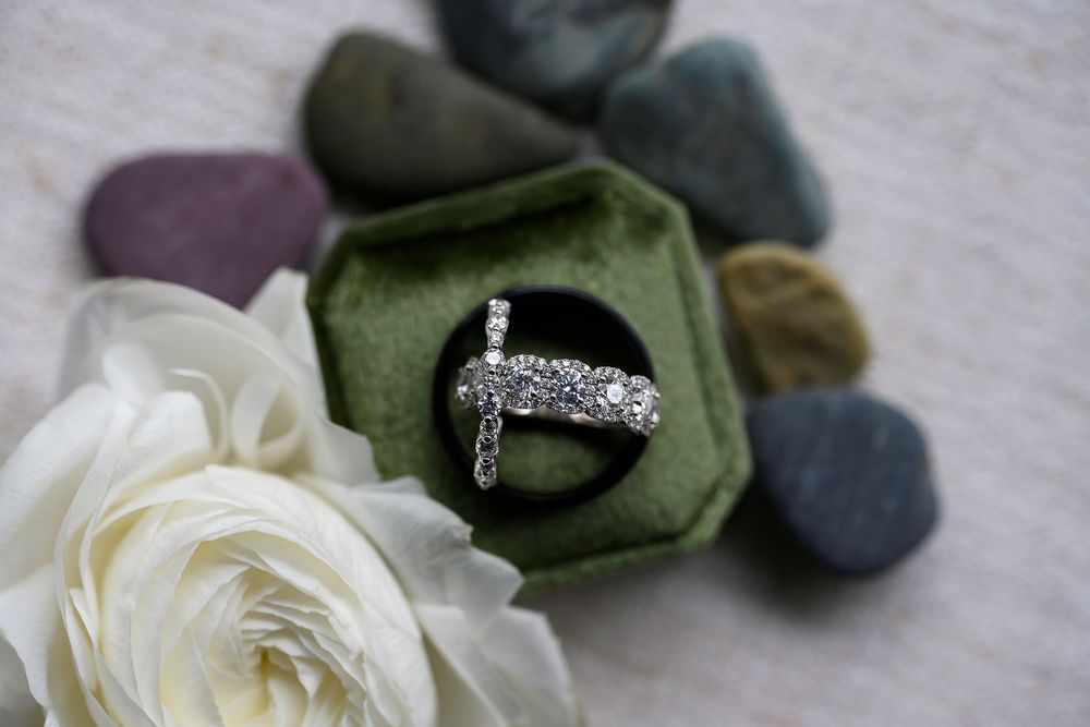 Diamond cross ring displayed in a green velvet ring box, surrounded by smooth stones and a white rose.