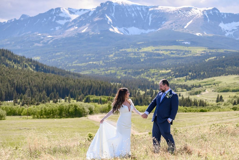 Bride and groom hold hands in a mountain meadow with snow-capped peaks and pine forests backdrop on their wedding day.