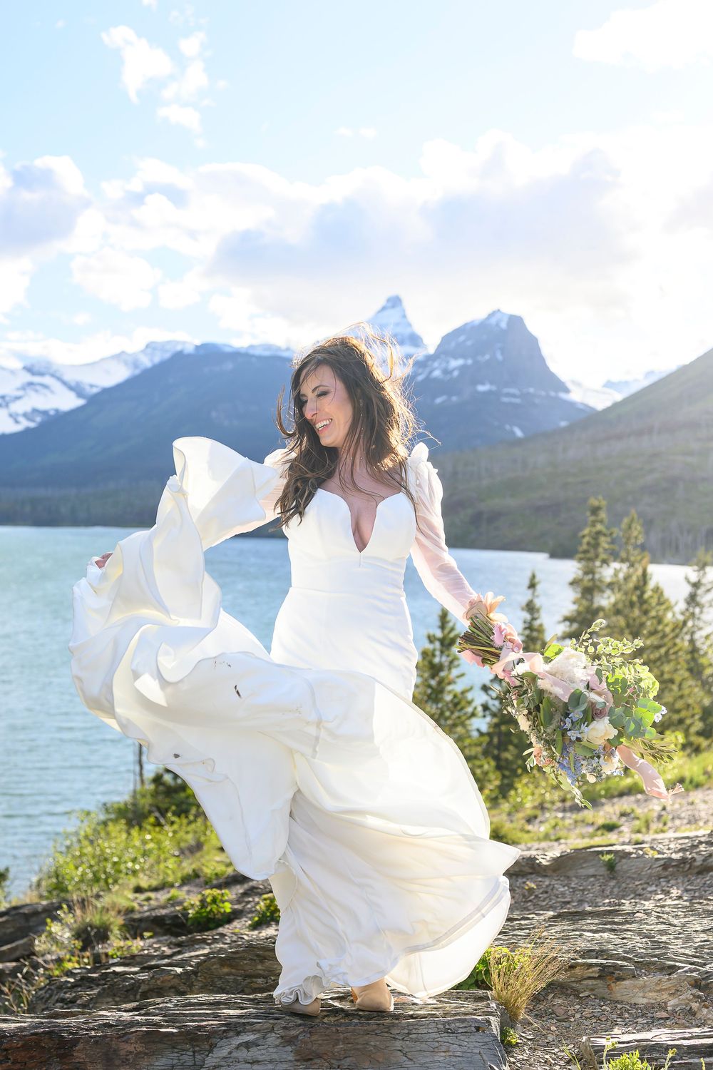 Bride in flowing white gown twirling by a mountain lake with snow-capped peaks and evergreen trees backdrop.