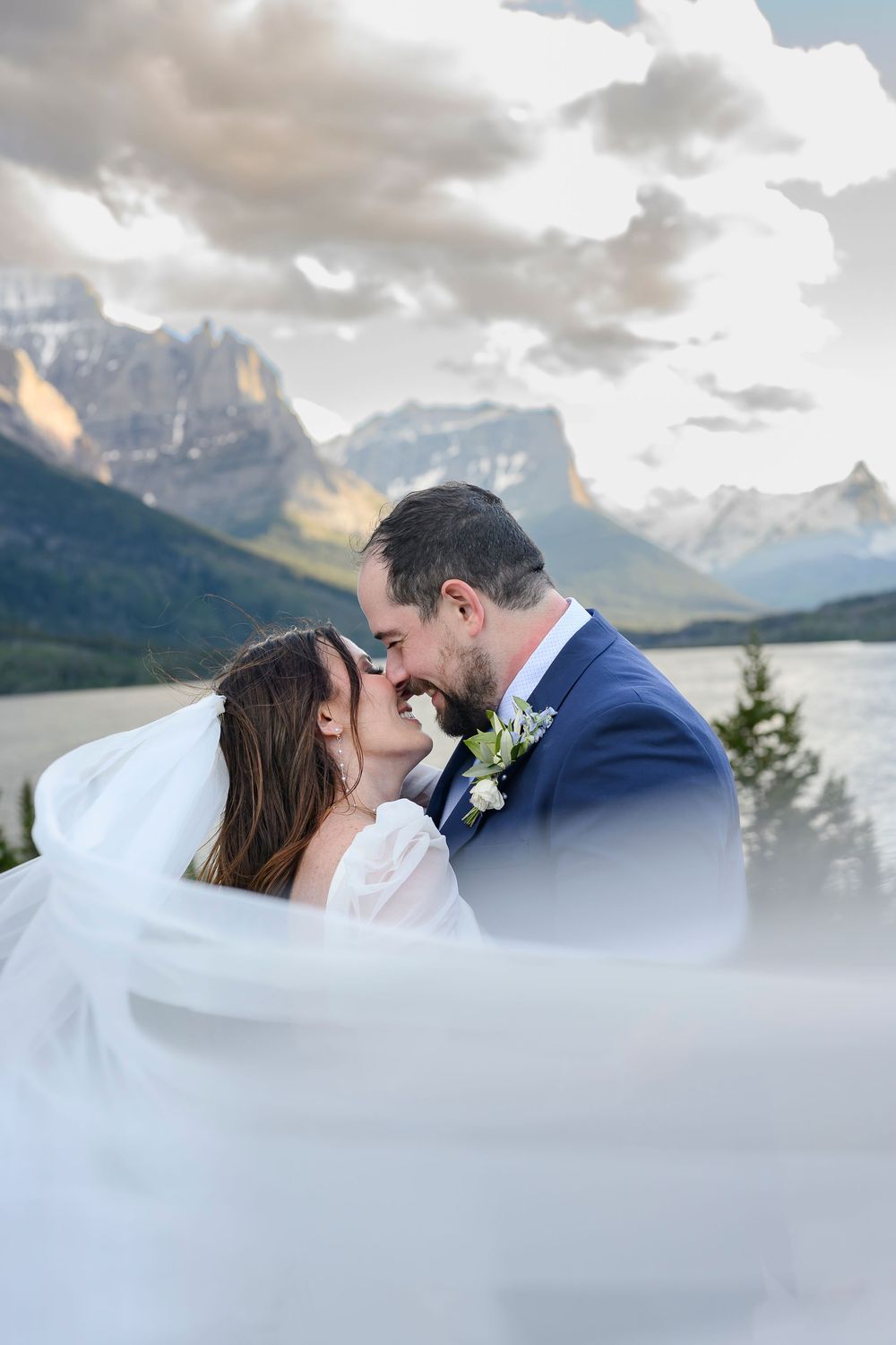 Bride and groom share a romantic kiss with veil flowing, set against stunning mountain lake backdrop.