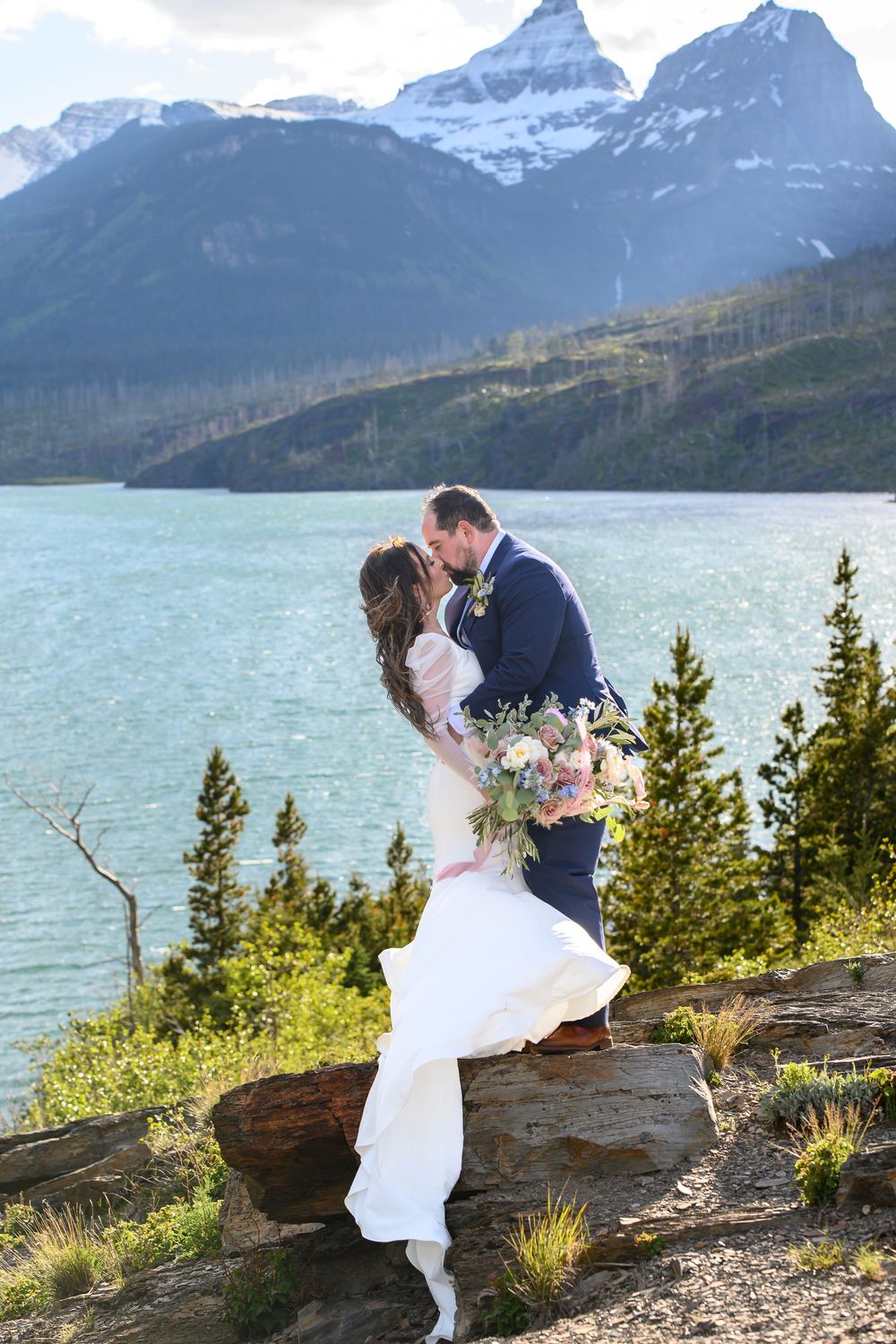 Bride and groom kissing on rocky overlook with mountain lake and snow-capped peaks backdrop at outdoor wedding.