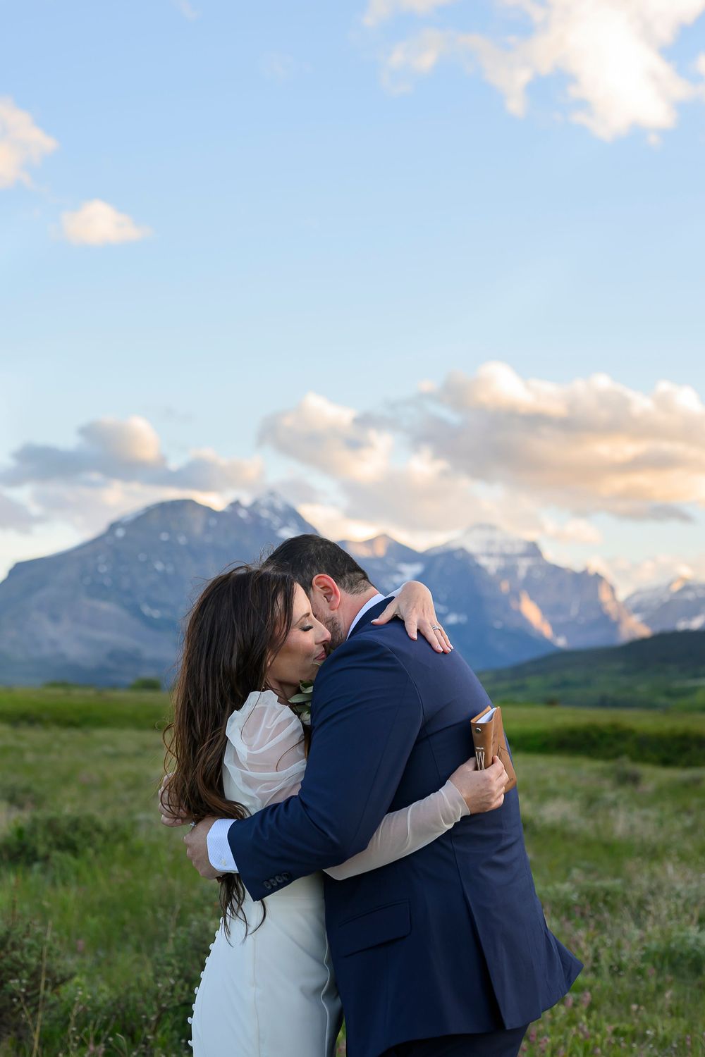 Bride and groom embracing in a green meadow with majestic mountain peaks and blue sky in the background.
