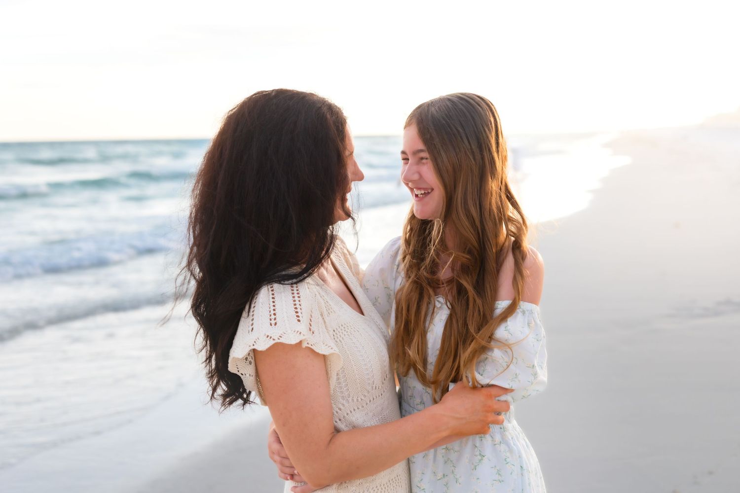 Two women in white dresses embrace and smile at each other on a sunny beach with ocean waves in the background.