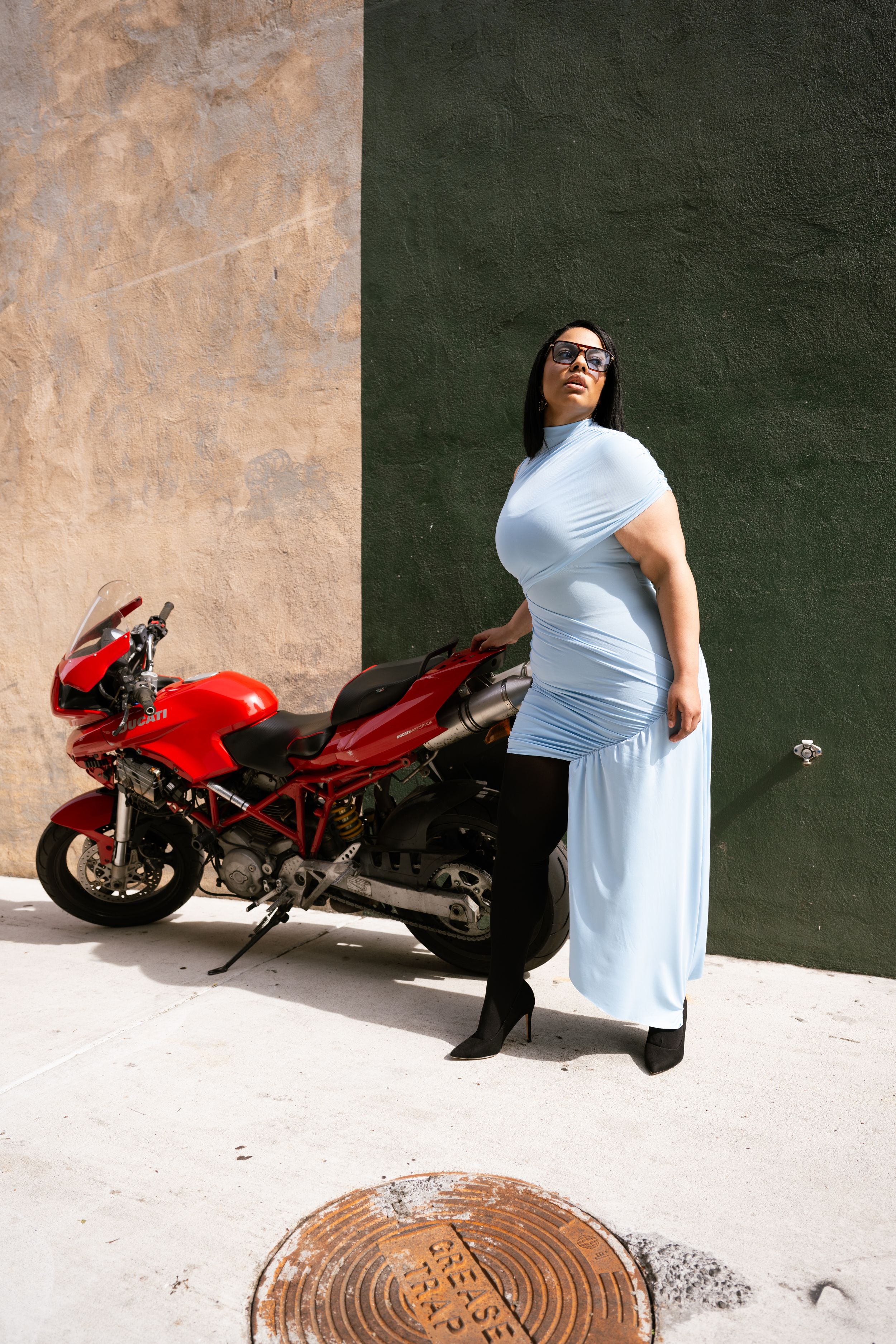 Woman in light blue dress and boots poses beside a red Ducati motorcycle against a green wall.